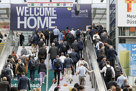A large crowd of people ascending escalators toward a sign that reads ‘WELCOME HOME’ at the Interpack event entrance.